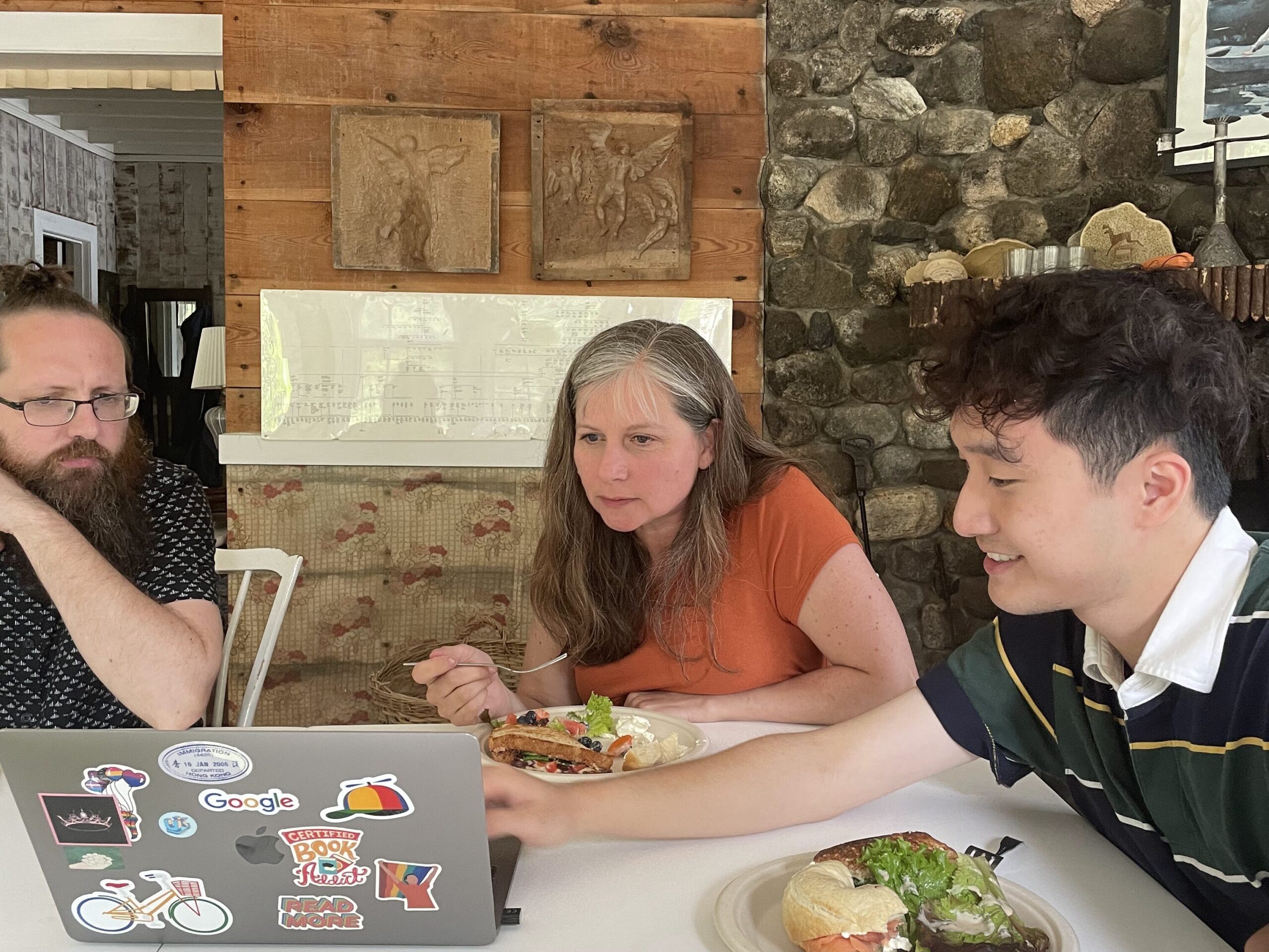 Three people sitting at a table eating food and looking at a computer.
