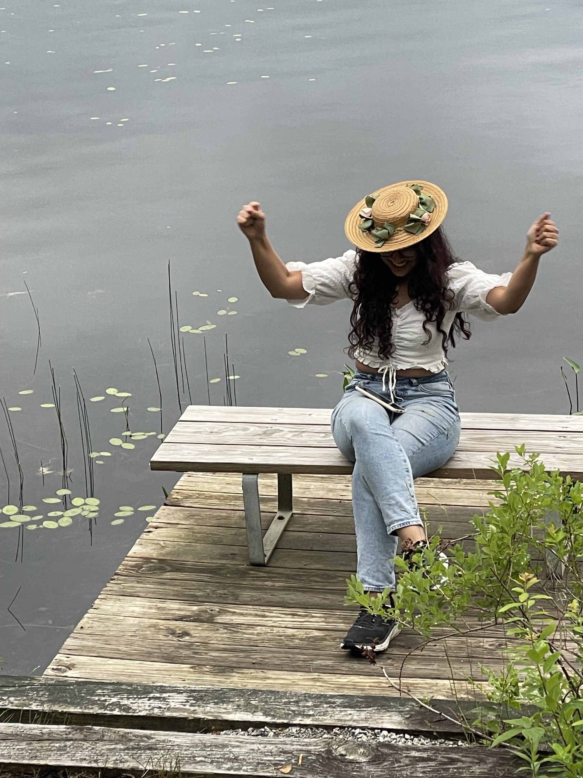 A person sitting on a bench near water holding their hands up and wearing a hat.